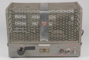 Audio instrument, amplifier. Rectangular, grey metal deivice with grate-like top with a silver strip down the center. Black dial, red and black buttons and switch.